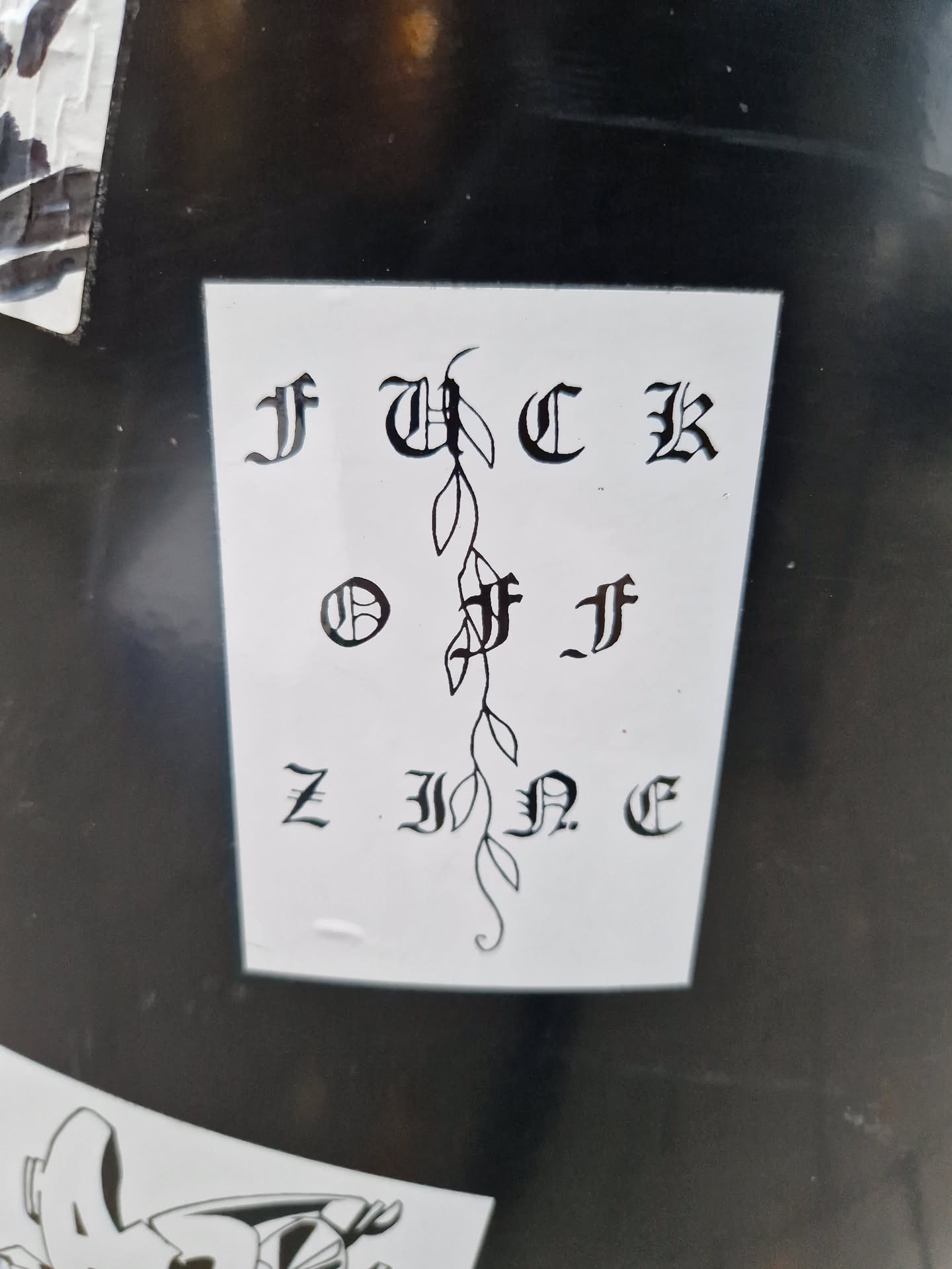 Black and white sticker with the text "FUCK OFF ZINE" written in a gothic style font. A small vine is featured in the center of the text.