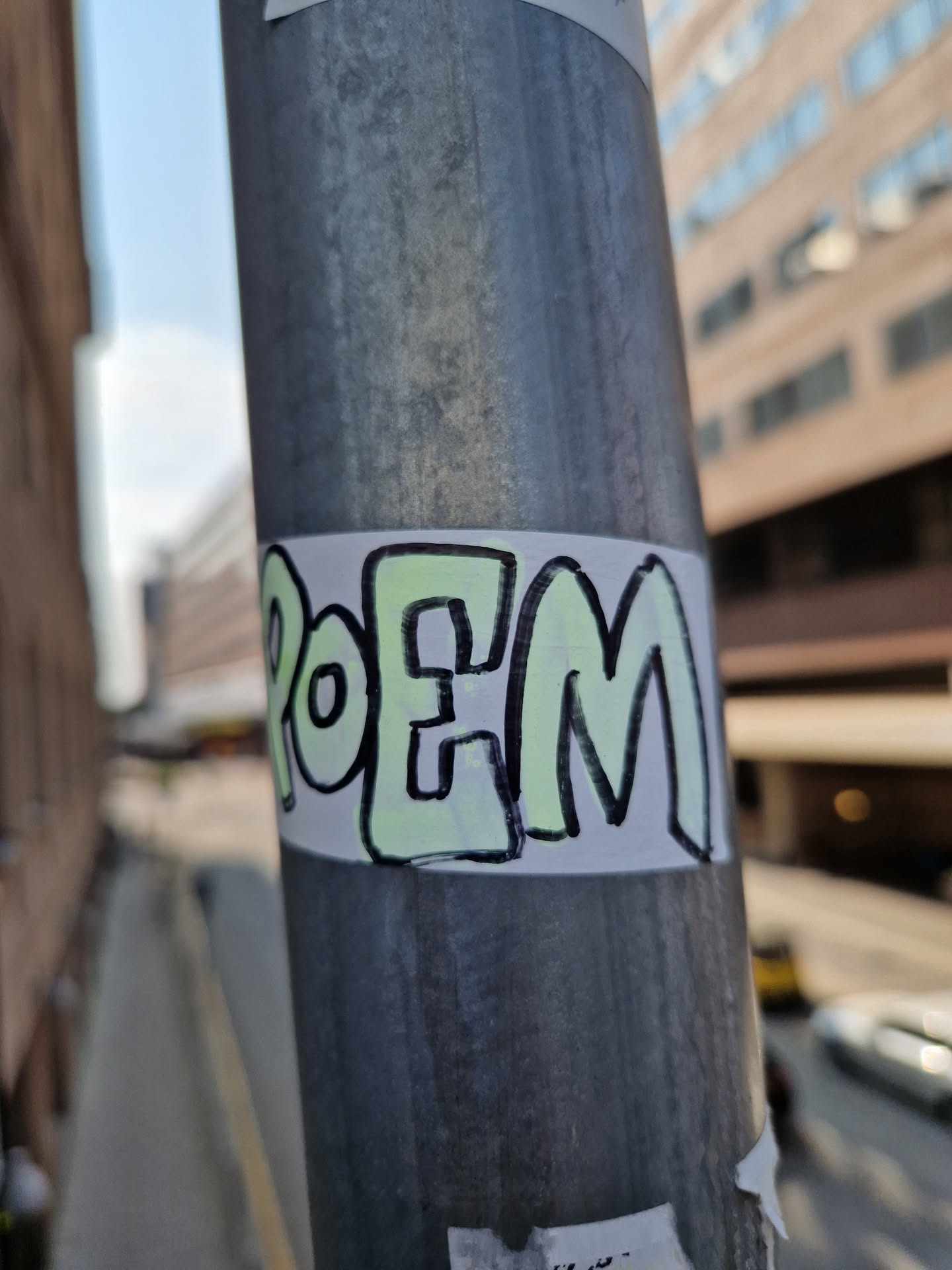 A light green and black sticker with the word POEM written in a stylized font. The sticker is affixed to a metallic pole outdoors.