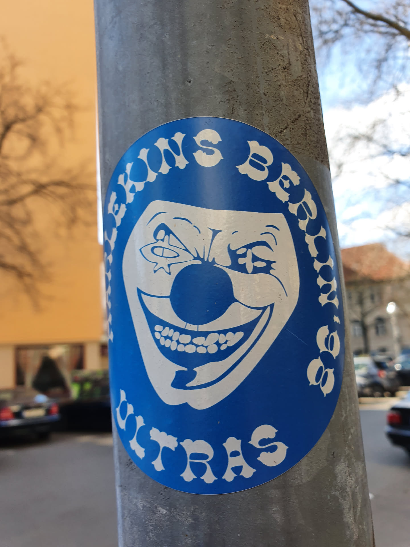 A blue circular sticker featuring a grinning clown face with unsettling details. The text around the image seems to read "LEBONS BERLIN 999 UTRAS". The style is reminiscent of vintage or retro designs, creating a unique and slightly disturbing aesthetic. The sticker is affixed to a metal pole in an urban environment.