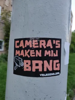 A sticker with a black background and peach colored text. It says 'Camera's Maken Mij Bang' which translates to 'Cameras Make Me Scared' in Dutch. There is a drawing of a security camera next to the text. At the bottom, there is a website address: vrijebond.org
