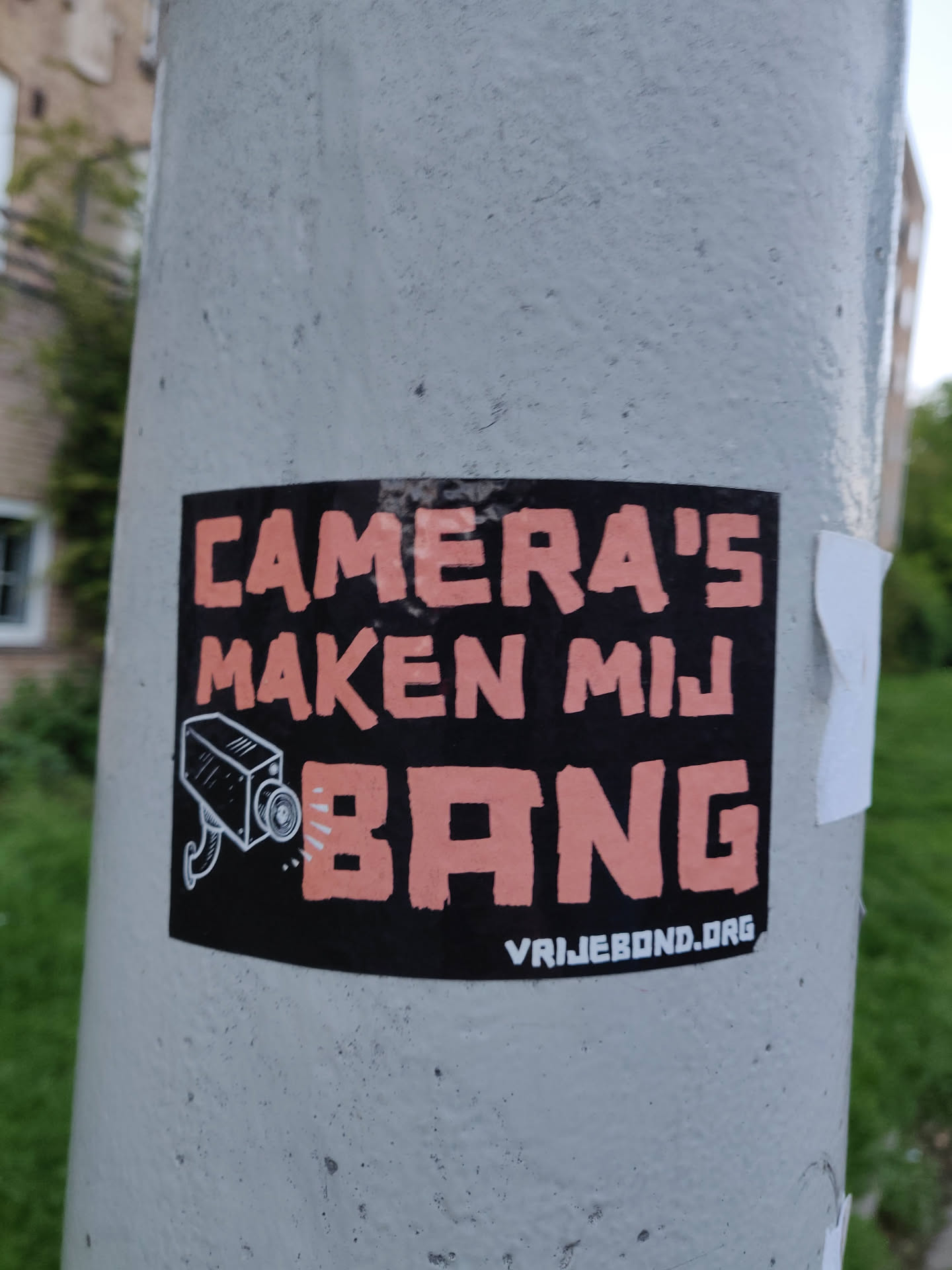 A sticker with a black background and peach colored text. It says 'Camera's Maken Mij Bang' which translates to 'Cameras Make Me Scared' in Dutch. There is a drawing of a security camera next to the text. At the bottom, there is a website address: vrijebond.org