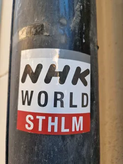 A sticker featuring the NHK WORLD logo and the abbreviation STHLM (Stockholm). The sticker is attached to a dark-colored pole outdoors.