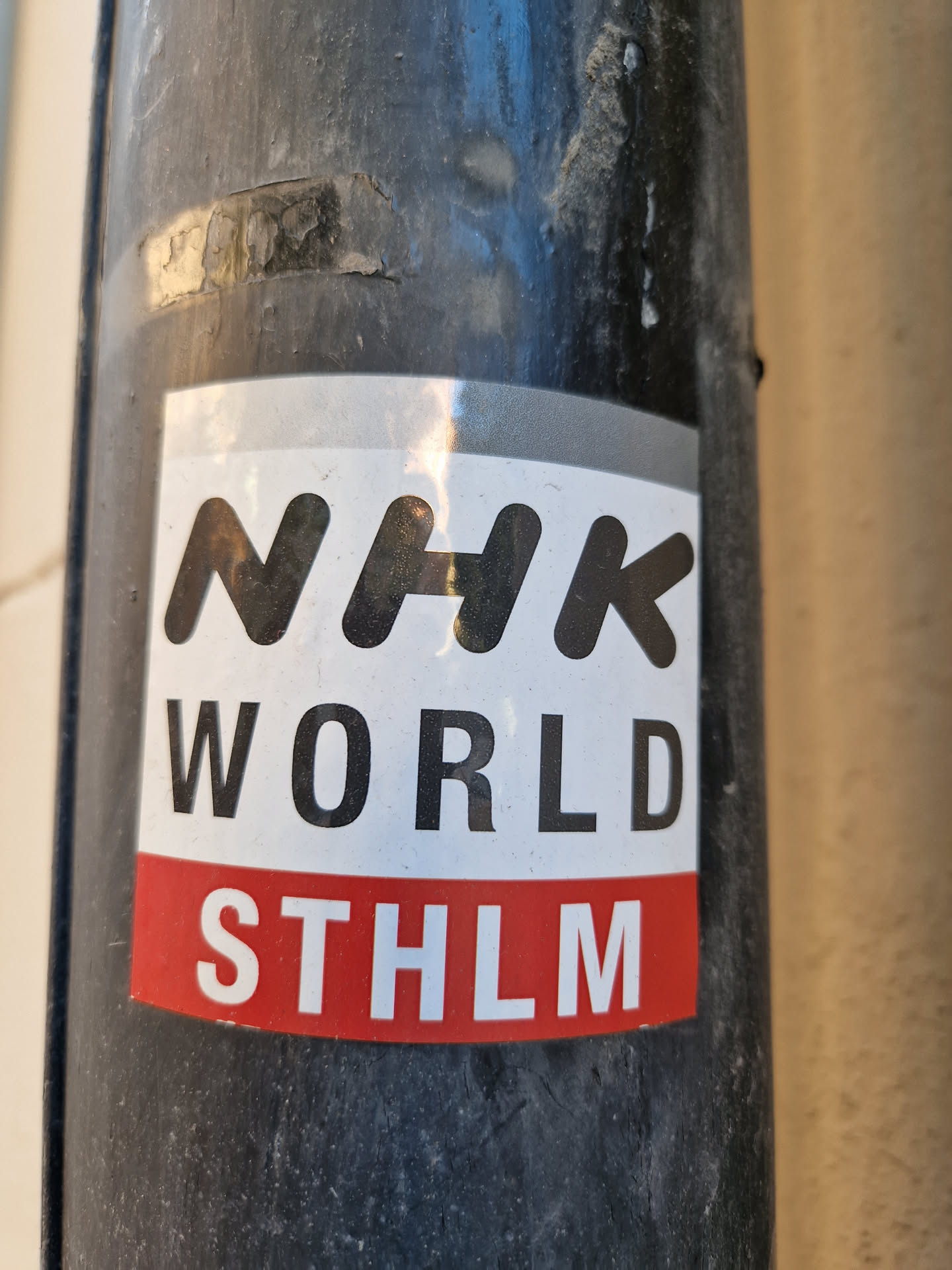 A sticker featuring the NHK WORLD logo and the abbreviation STHLM (Stockholm). The sticker is attached to a dark-colored pole outdoors.