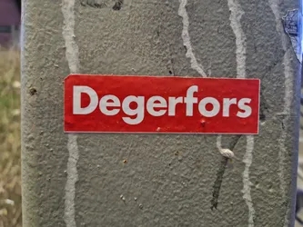 A red sticker with the word Degerfors in white letters is affixed to a grey pole. The pole shows signs of wear and other markings.
