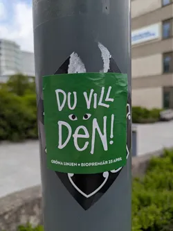 A square, slightly crumpled green sticker attached to a gray pole. The sticker features the text "Du Vill Den!" in bold white letters, with two small circles above the text that resemble eyes. Below the main text, smaller text reads "GRÖNA LINJEN • BIOPREMIÄR 25 APRIL".  The sticker is adhered to a gray pole, partly covering another black and white sticker with a diamond and curved line design.