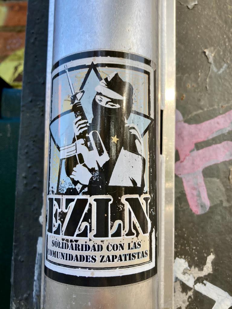 The sticker is primarily black and white, with some areas appearing light blue or faded yellow due to lighting and wear. It depicts a central, stylized figure, likely a Zapatista rebel, wearing a balaclava or ski mask that obscures their face, along with a wide-brimmed hat. The figure holds a rifle and has a small, five-pointed star visible on their chest. Behind the figure, there's a large, distressed five-pointed star design. Below the main image, the bold, capitalized letters "EZLN" are prominently displayed. Underneath "EZLN," in smaller, capitalized Spanish text, it reads "SOLIDARIDAD CON LAS COMUNIDADES ZAPATISTAS." The overall aesthetic is stark and graphic, reminiscent of stencil art, applied to a silver pole.