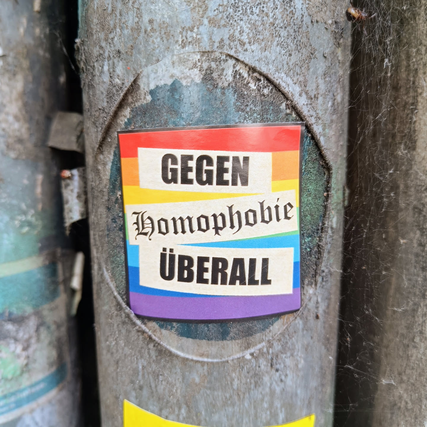 A sticker with a rainbow flag background and the text "Gegen Homophobie überall" which translates to "Against Homophobia everywhere" in German. The sticker is attached to a metal pole.