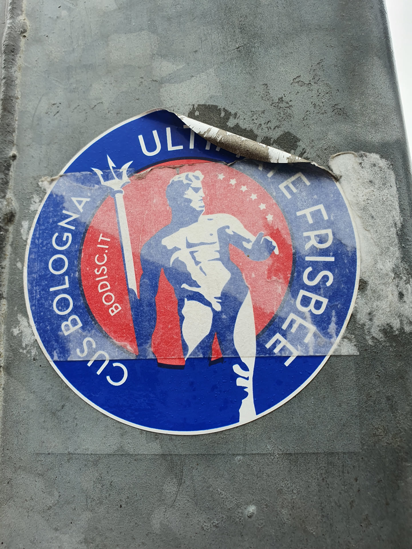 A worn sticker featuring a circular design with a figure holding a trident. The text includes "Ultimate Frisbee" and "Bologna". The color scheme is predominantly blue and red.