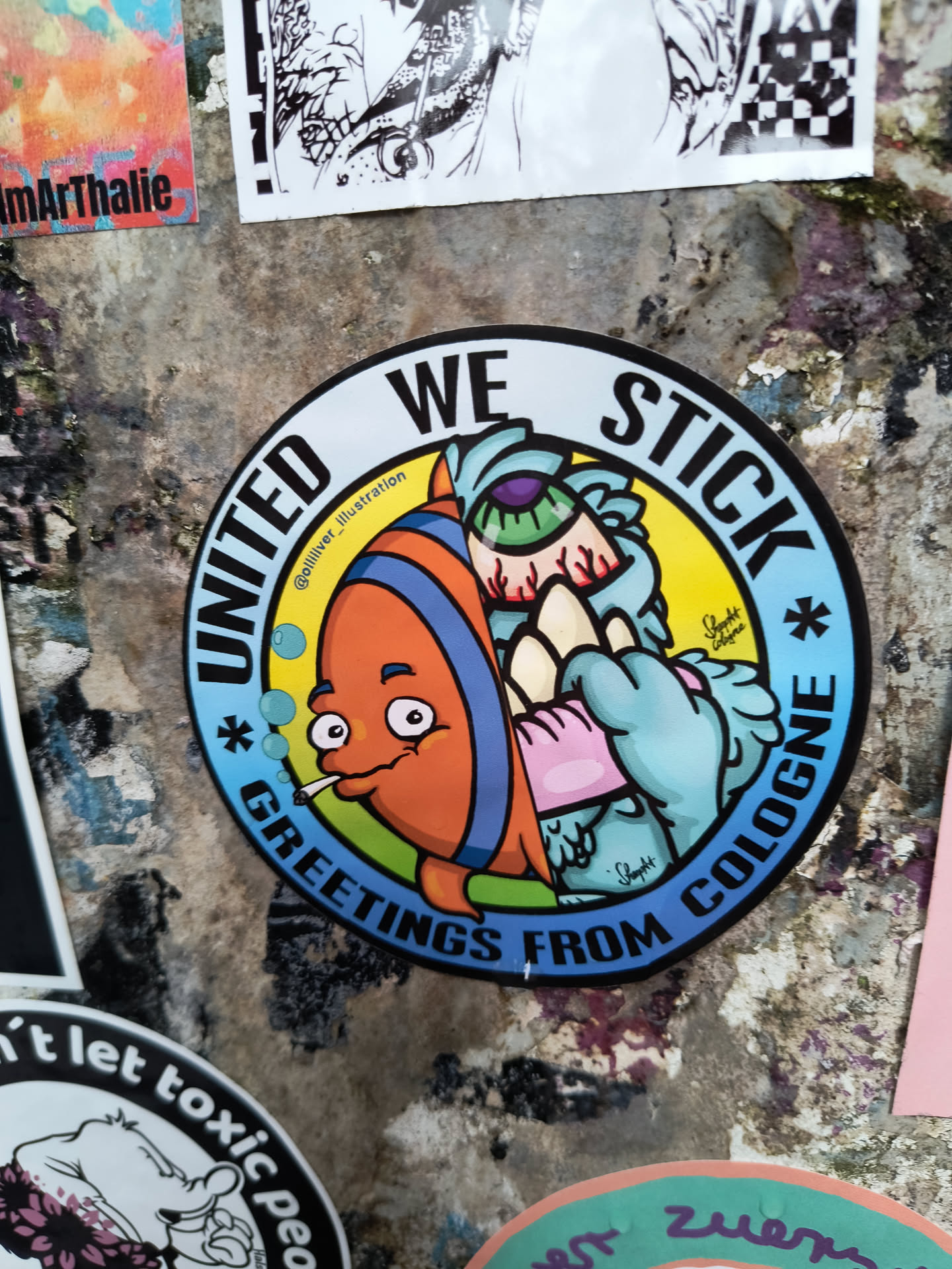 This round sticker shows a cartoon fish smoking and a light blue monster in the middle. The text says "UNITED WE STICK * GREETINGS FROM COLOGNE *" around the edge of the sticker.