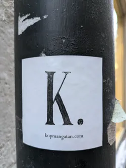 A square white sticker with the letter "K" and a small period to the right of it, followed by "kopmangatan.com" at the bottom. It's affixed to a black pole.