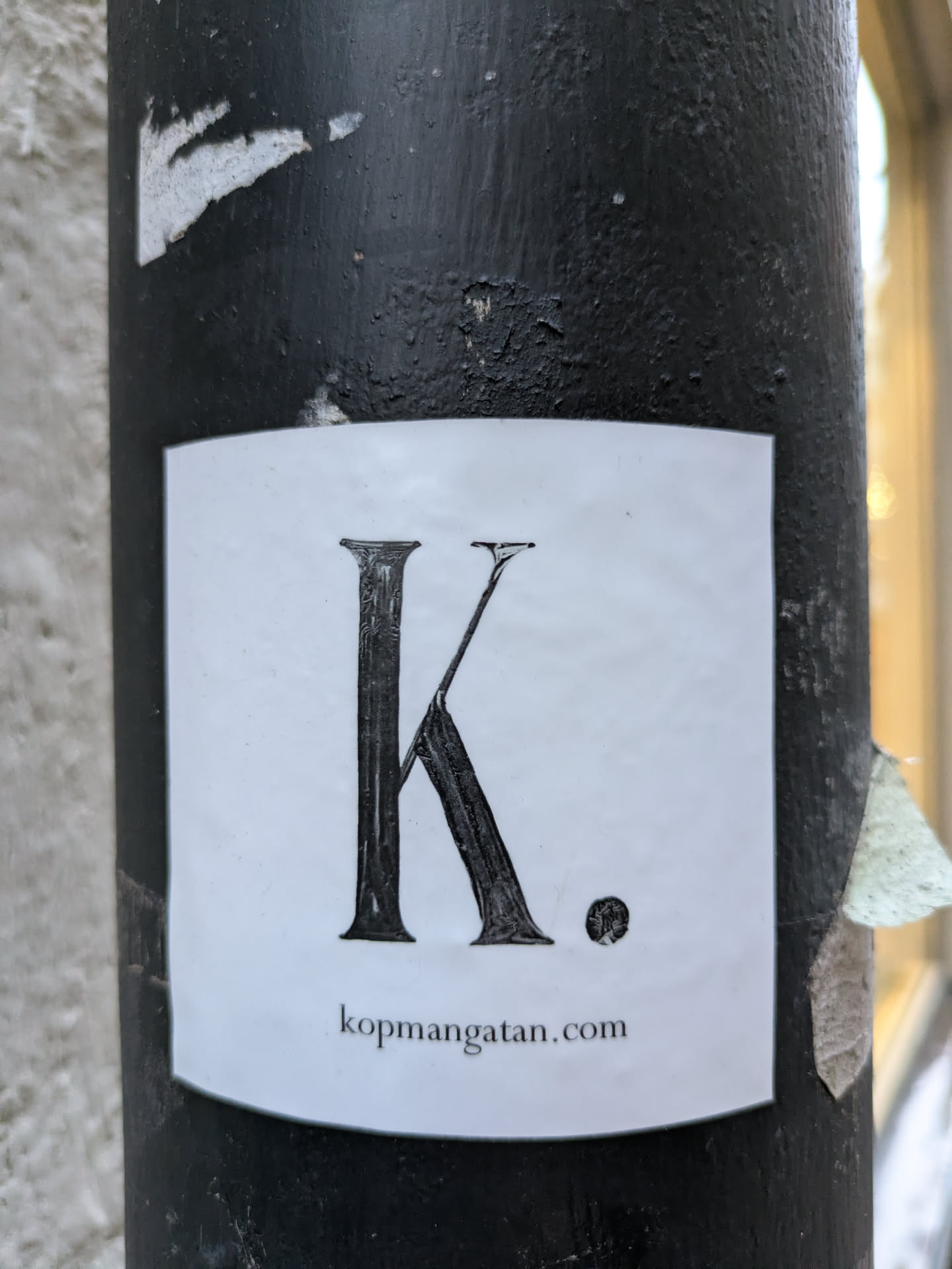 A square white sticker with the letter "K" and a small period to the right of it, followed by "kopmangatan.com" at the bottom. It's affixed to a black pole.