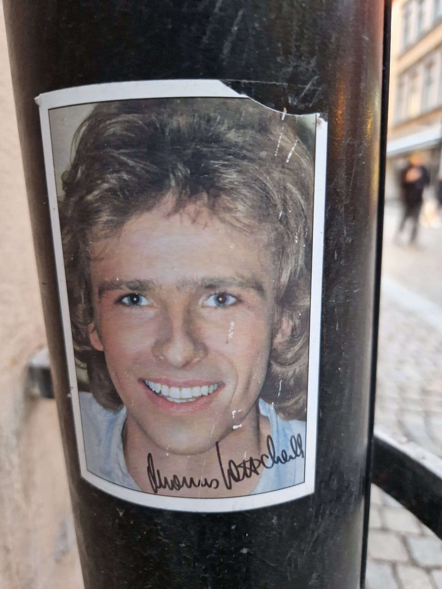 A sticker featuring a portrait of British actor Dennis Waterman, with his signature underneath. The sticker is affixed to a dark pole outdoors.