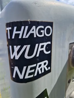A black and white sticker with the text "THIAGO WUFC NERR." on it. The sticker is affixed to a gray surface outdoors.