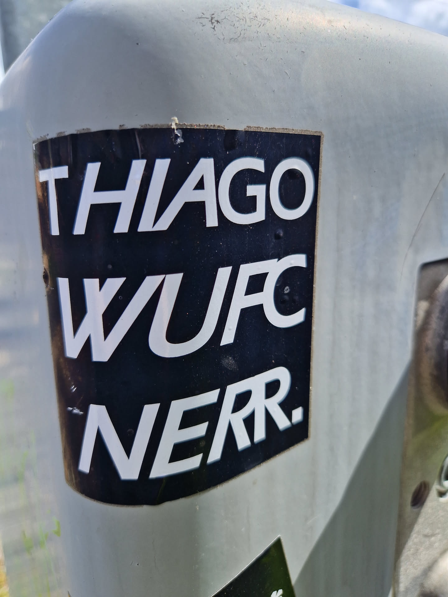 A black and white sticker with the text "THIAGO WUFC NERR." on it. The sticker is affixed to a gray surface outdoors.