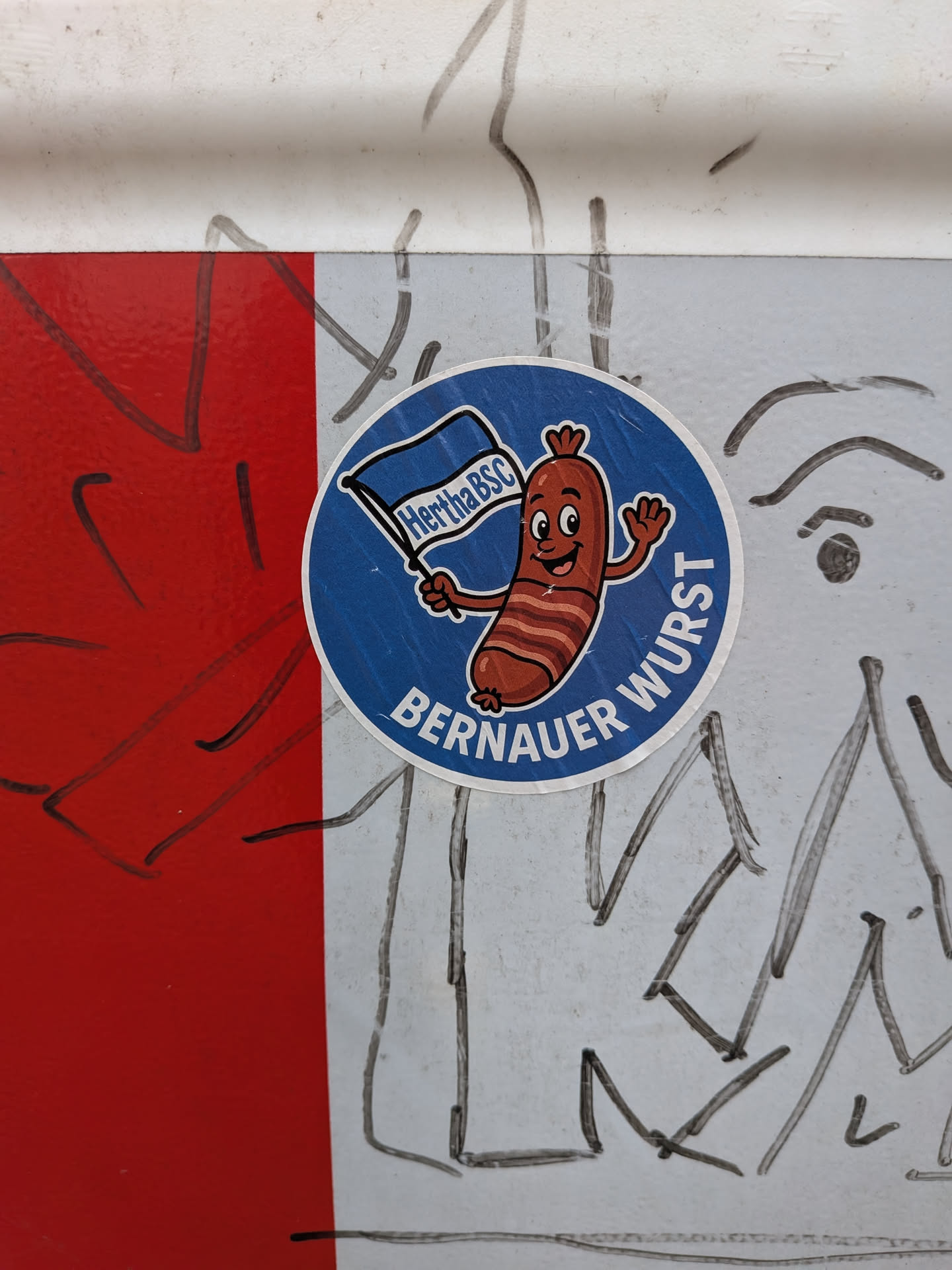 A circular sticker set against a solid blue background framed by a thin white border. At the center is a cheerful cartoon sausage mascot with grill marks, waving a blue-and-white Hertha BSC flag with one hand and waving hello with the other. Below the mascot, the words 'BERNAUER WURST' are printed in an arched, bold white sans-serif font.