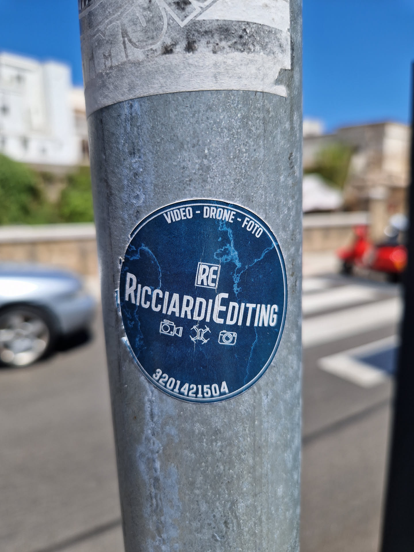 A blue round sticker with the text RicciardieEditing, Video - Drone - Foto, and the number 3201421504. It features icons for a camera, a drone, and a video camera. The sticker is affixed to a metal pole.