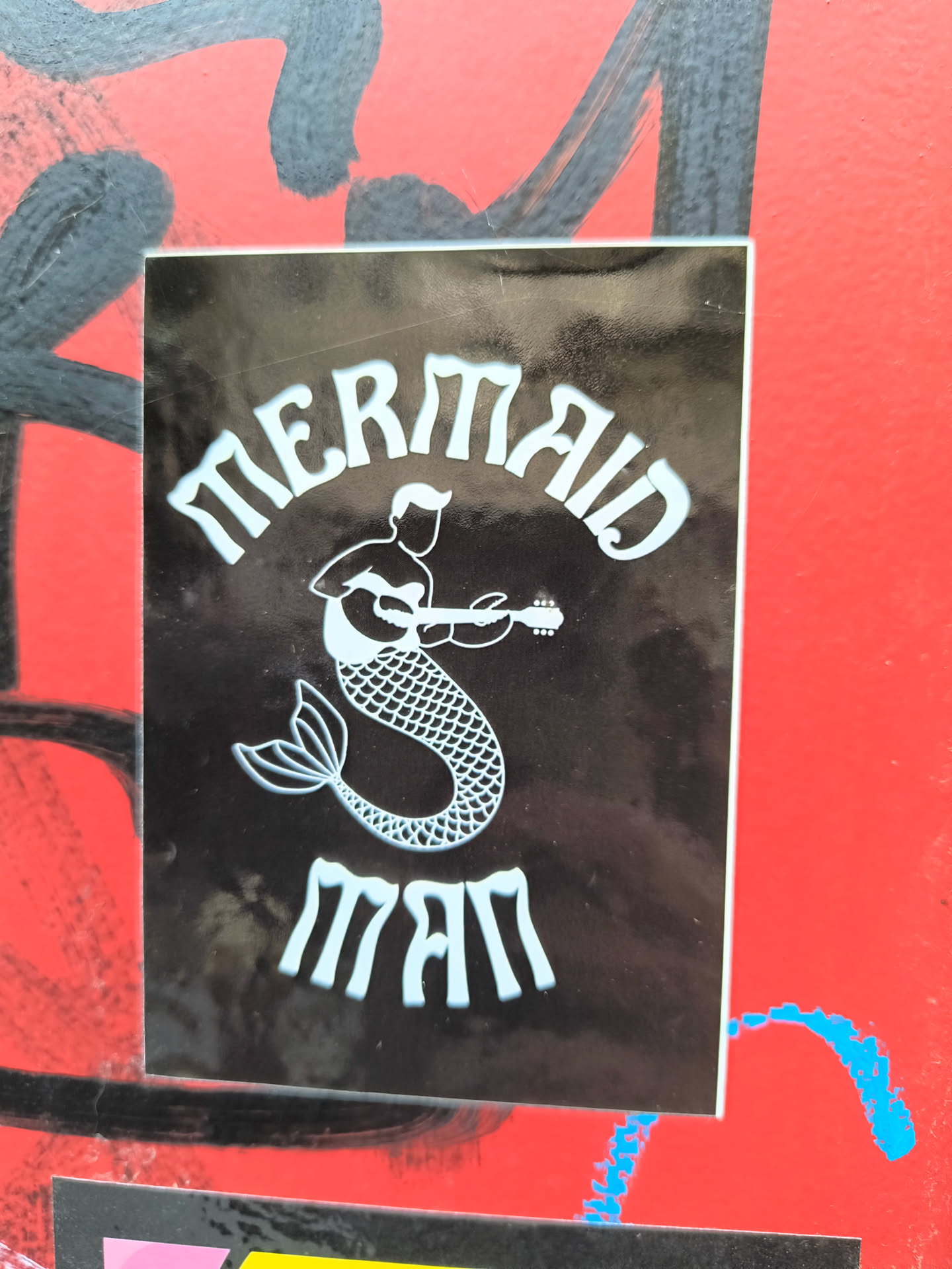 The sticker is a rectangular, vertically oriented design with a glossy black background. It features a stylized illustration in a light blue or off-white outline. The central image is a male figure, a merman, depicted from the waist up as human with a distinct hairstyle, playing an acoustic guitar. His lower body transitions into a detailed fish tail with visible scales and a classic fluke. Above the merman, curved in an ornate, slightly blocky font, is the word "MERMAID". Below the merman, in the same font, is the word "MAN". The overall appearance is clean and graphic, with a subtle sheen on its surface, indicating a likely laminated finish. The sticker appears adhered to a red, graffiti-covered wall, showing slight wear and reflections.