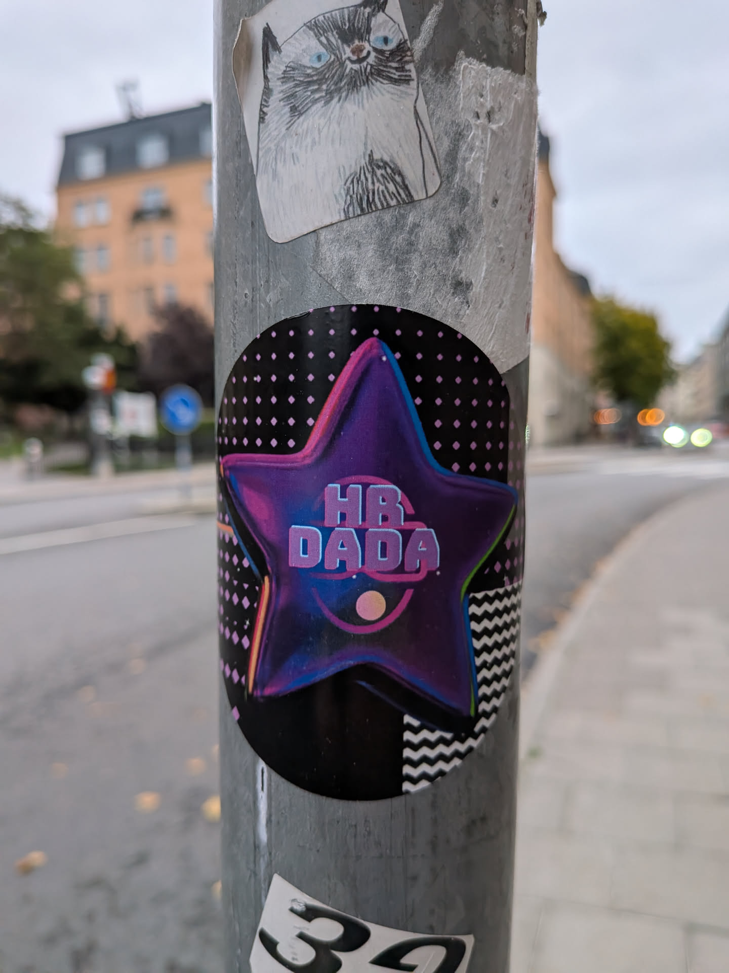 An oval sticker featuring a central, rounded five-pointed star in deep purple with glowing neon pink and cyan outlines. In the center of the star, retro-futuristic block lettering spells out 'HR DADA' in light purple, accompanied by a small circular graphic and an arc underneath. The dark background incorporates retro graphic elements, including a grid of pink dots and a black-and-white zigzag chevron pattern in the bottom right corner.