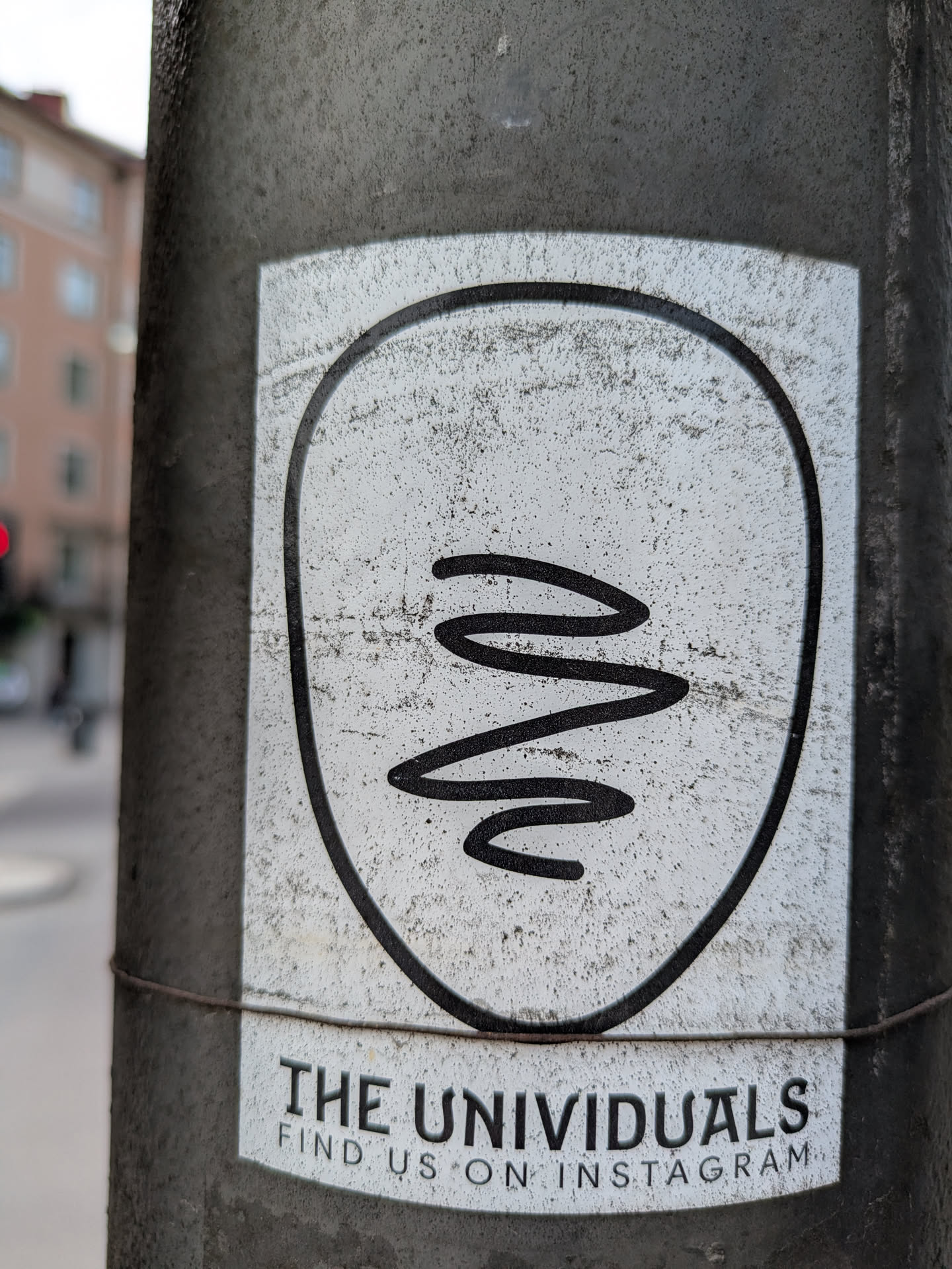 This sticker on a pole features a minimalist design with a squiggly line inside a rounded frame. Below the design, the text 'THE UNIVIDUALS' is printed, followed by 'FIND US ON INSTAGRAM', indicating a social media presence.