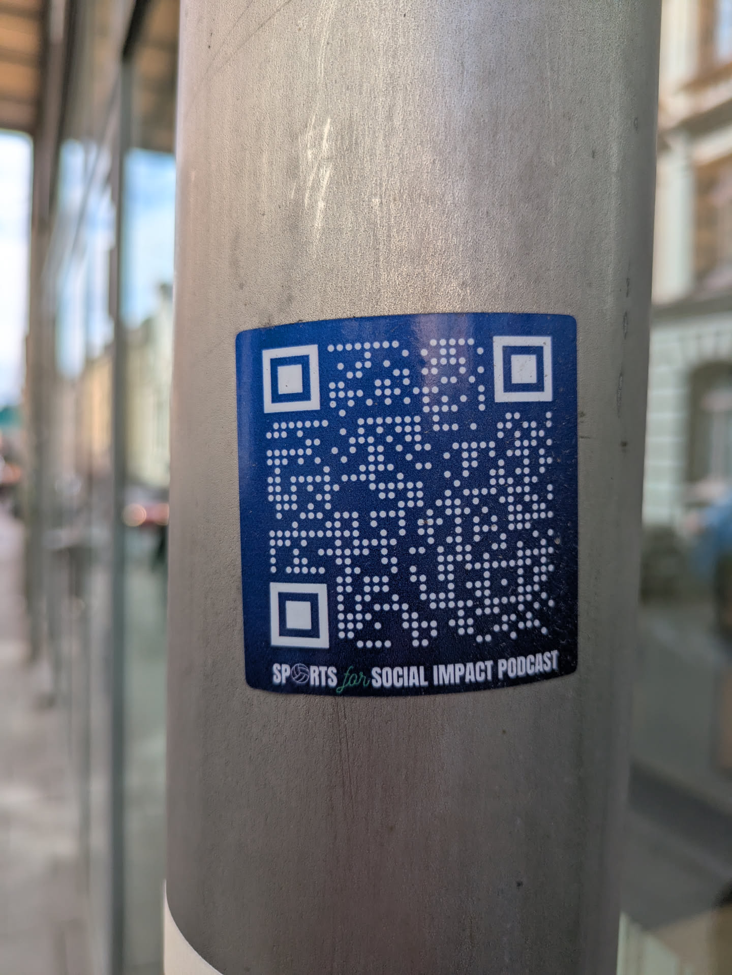 This sticker is attached to a light grey metallic pole and features a blue background with a white QR code. At the bottom is the text 'SPORTS for SOCIAL IMPACT PODCAST'. The QR code likely leads to the podcast's website or social media page.