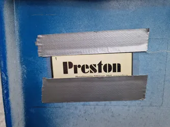 A close-up shot of a sticker featuring the word "Preston" in bold, uppercase letters. The sticker is affixed to a blue surface using gray duct tape.  Small text at the bottom of the sticker is partially visible.