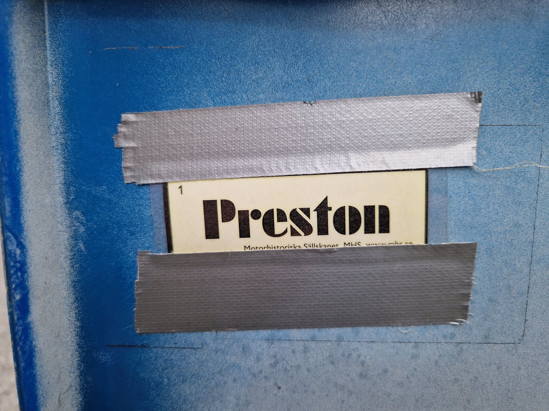 A close-up shot of a sticker featuring the word "Preston" in bold, uppercase letters. The sticker is affixed to a blue surface using gray duct tape.  Small text at the bottom of the sticker is partially visible.