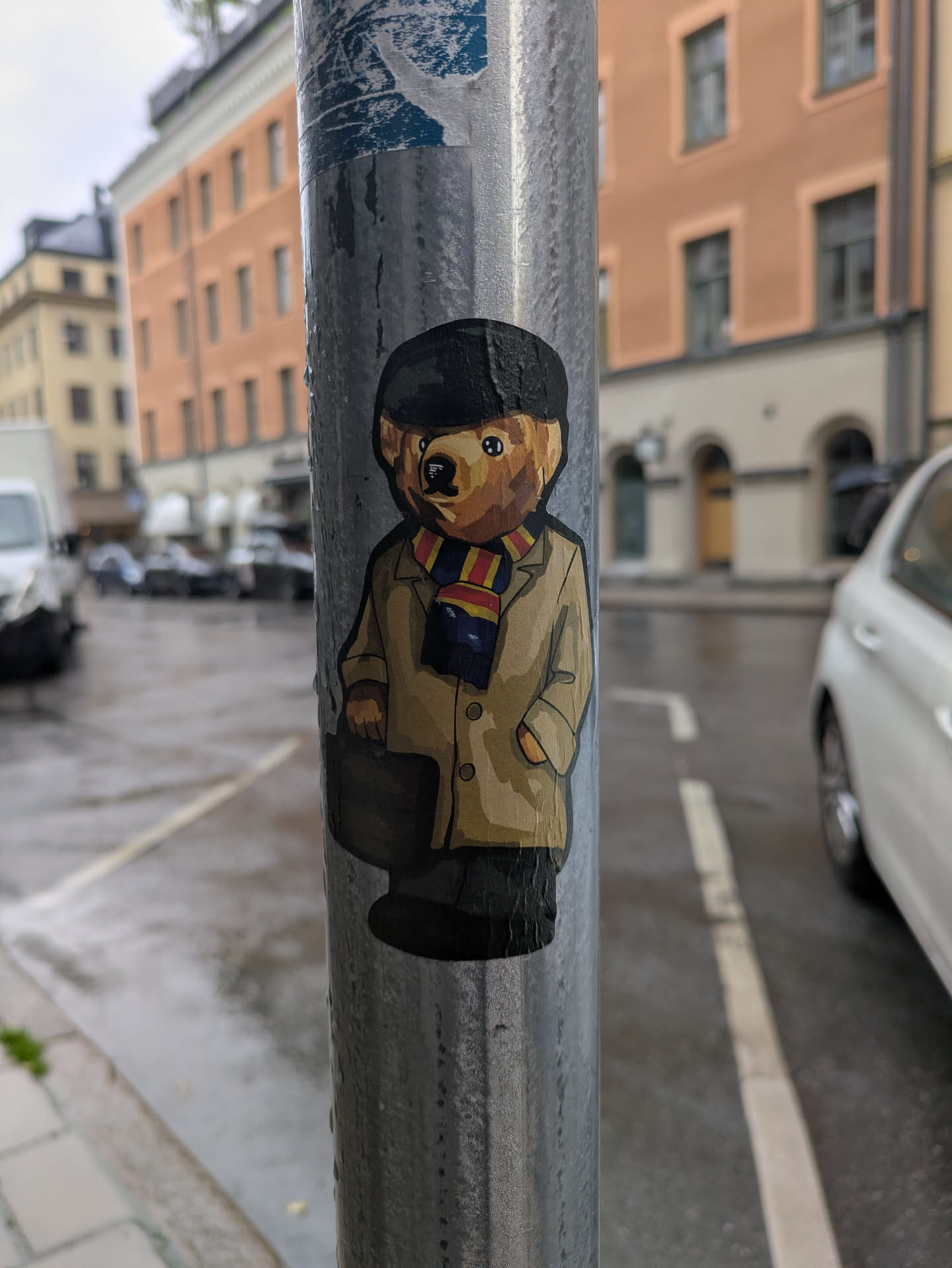 A sticker of a bear dressed in a cap, coat, and scarf, holding a briefcase, is affixed to a metal pole. The bear gives off a professional and sophisticated vibe.