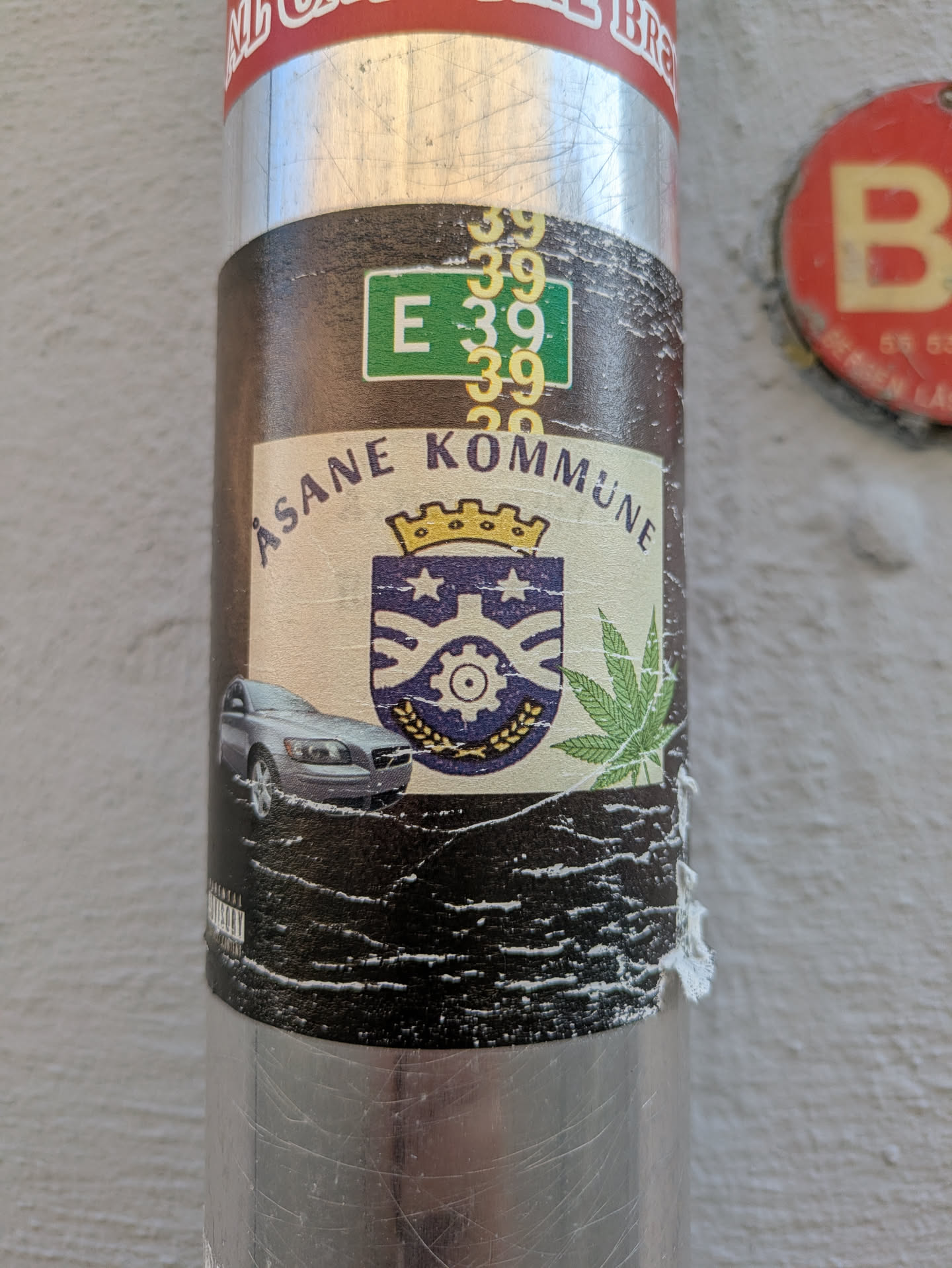 The sticker, affixed to a metal pole, features a central cream-colored banner with the text "ÅSANE KOMMUNE" curved above a blue and gold crest or coat of arms, topped with a crown. To the left of the crest is a silver Volvo car, and to the right is a prominent green cannabis leaf. Above the banner, a green rectangle displays "E 39", surrounded by several instances of the number "39" in yellow. The background of the sticker below the banner is dark brown or black, showing signs of wear and peeling.