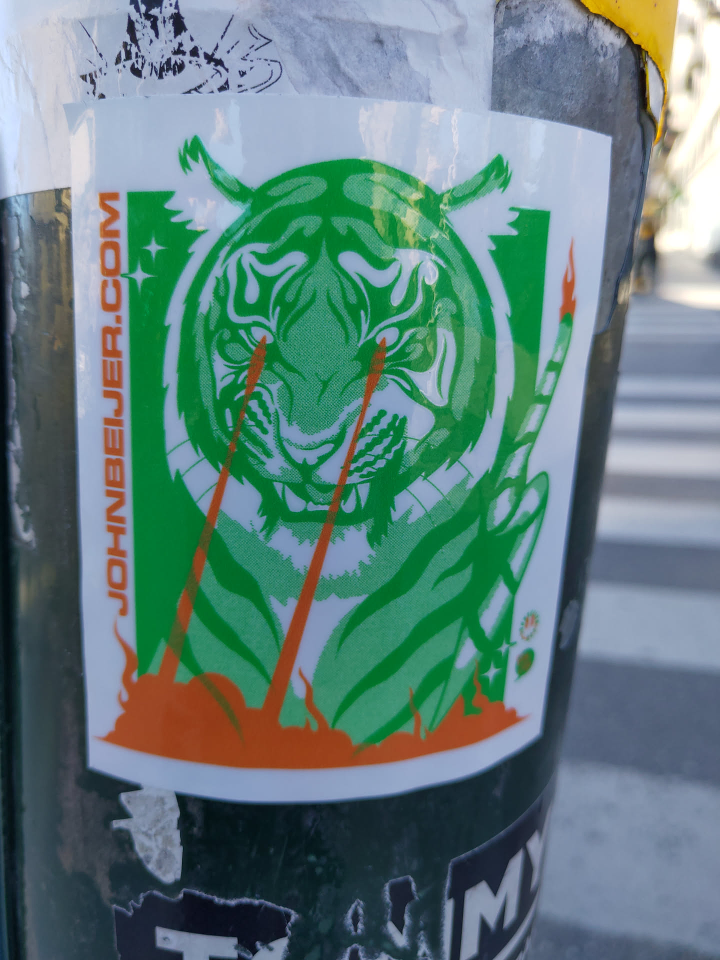 Green and orange sticker of a tiger shooting lasers from its eyes. The sticker has the website JOHNHEYER.COM printed on it. 