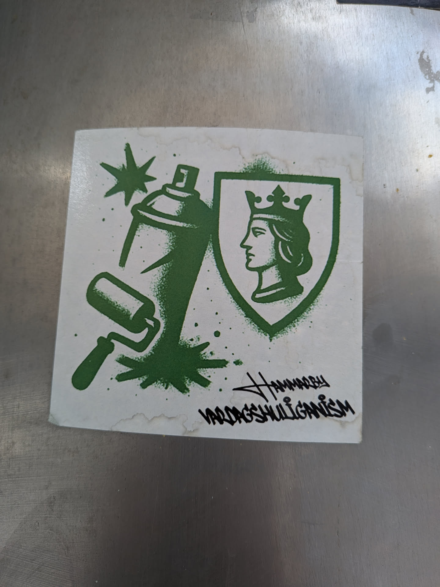 A square-shaped sticker with a white background and dark green imagery. On the left, a spray paint can is depicted, emitting a burst of paint with star-like elements and splatters. Below the can, a paint roller is shown. On the right, a shield emblem contains the profile of a crowned head, facing left. The green elements have a textured, spray-painted appearance. Below the main graphic, there are two lines of text in a stylized, graffiti-inspired font. The edges of the white sticker show some minor wear and light brown discoloration, suggesting it's a physical sticker that has been applied.