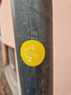 The sticker features a simple illustration of what seems to be two figures or objects in a balanced position, possibly referencing a seesaw or a similar concept. The style is clean and uncluttered. The sticker is affixed to a weathered, dark gray pole.