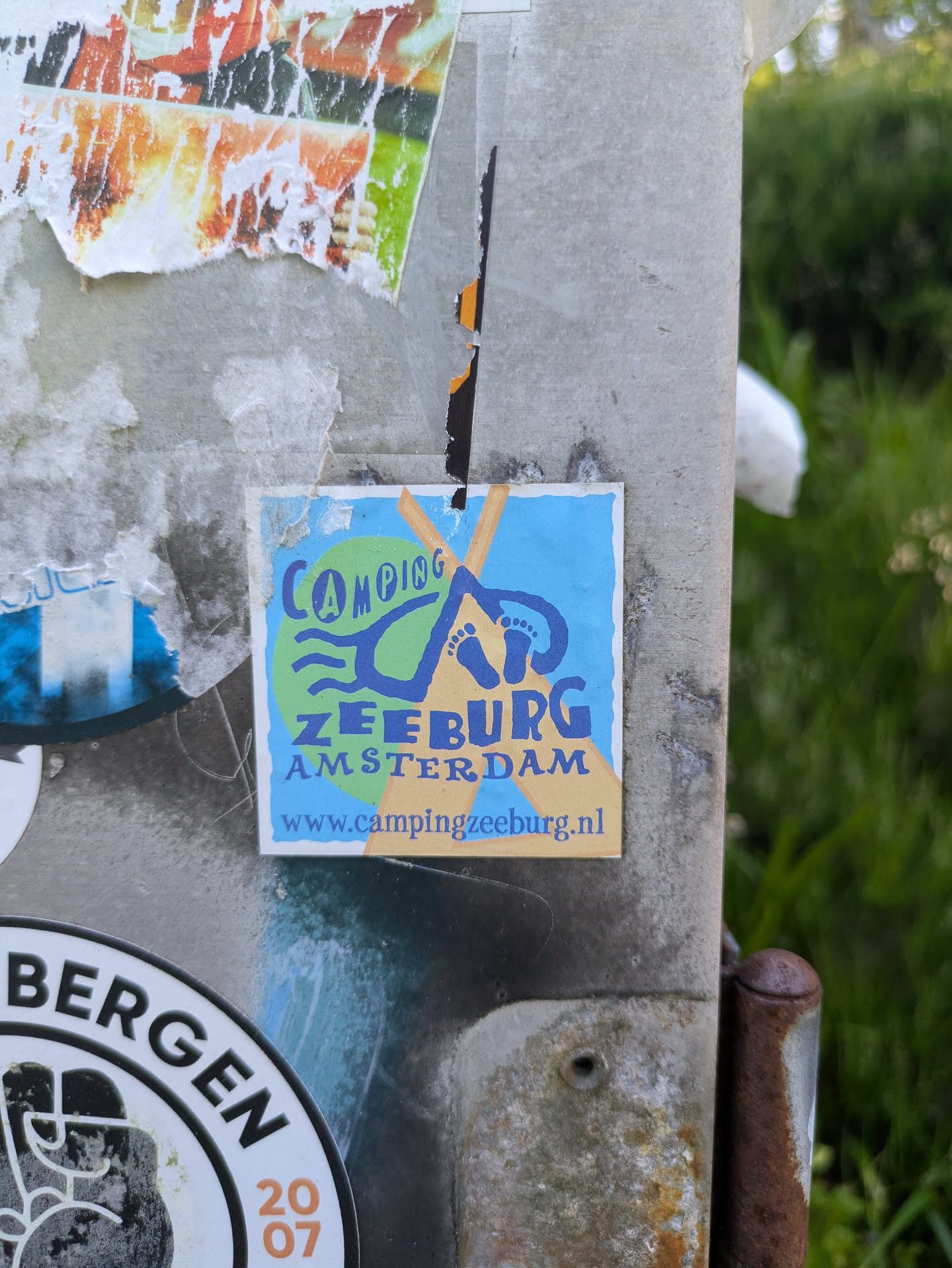 The central sticker is square and colorful, featuring a light blue background with a large green circle and orange-yellow cross-shaped elements. Stylized blue graphics depict waves, a tent, and a pair of footprints, signifying an outdoor or camping theme. Below the main graphic, the name of the campsite and city are clearly written. At the very bottom, a website address is provided. The sticker is slightly worn at the edges and is placed on a textured, grey metal pole that also shows remnants of other torn stickers and some rust, with green foliage visible in the blurred background.