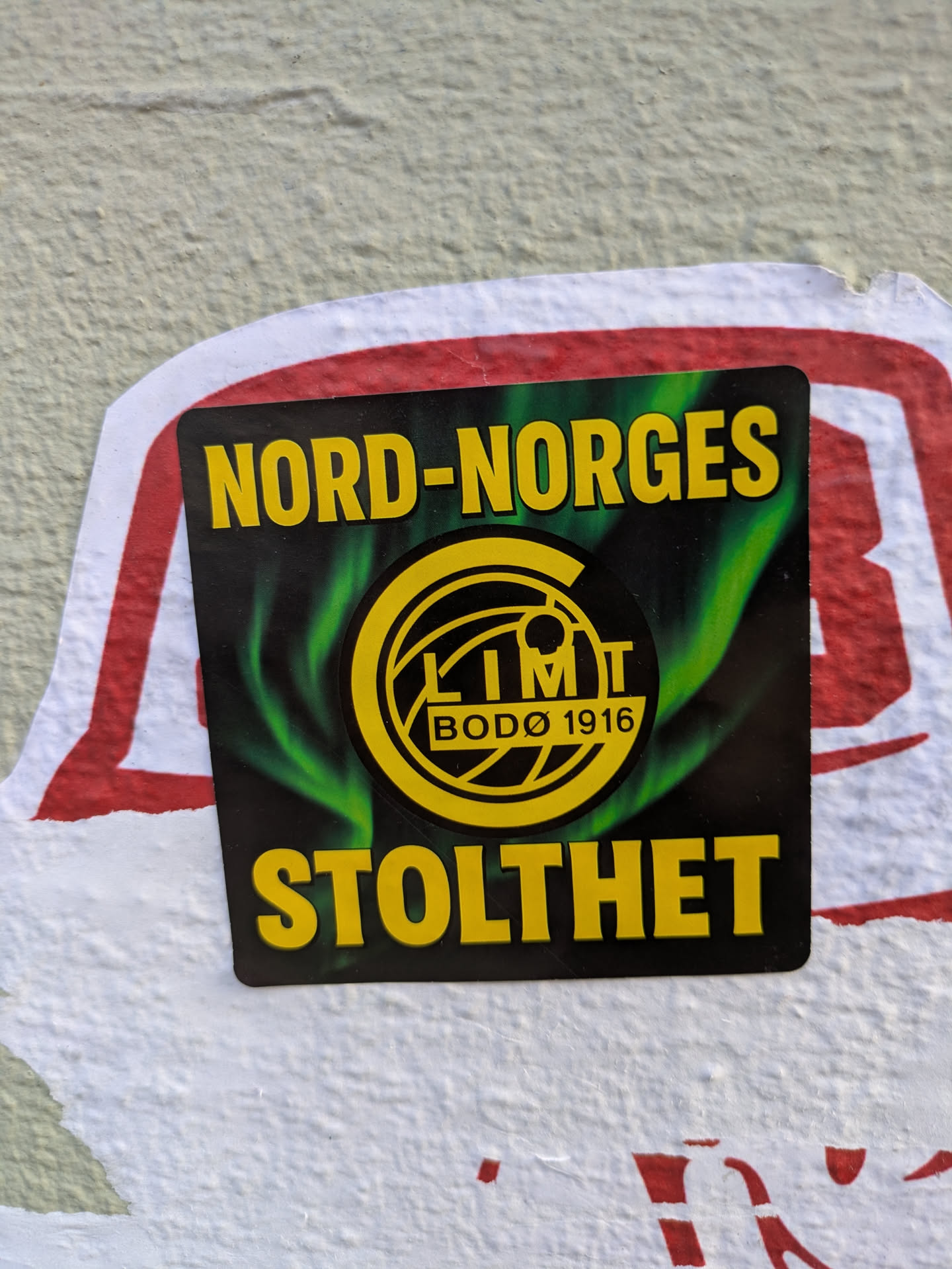 The sticker is rectangular, predominantly black with a vibrant green swirling pattern behind a central yellow circular logo. The logo contains the text "GLIMT" and "BODØ 1916". Above the logo, in large yellow capital letters, is "NORD-NORGES", and below it, also in large yellow capitals, is "STOLTHET". The sticker is applied to a light-colored, textured wall and partially overlaps a faded red and white sticker underneath.
