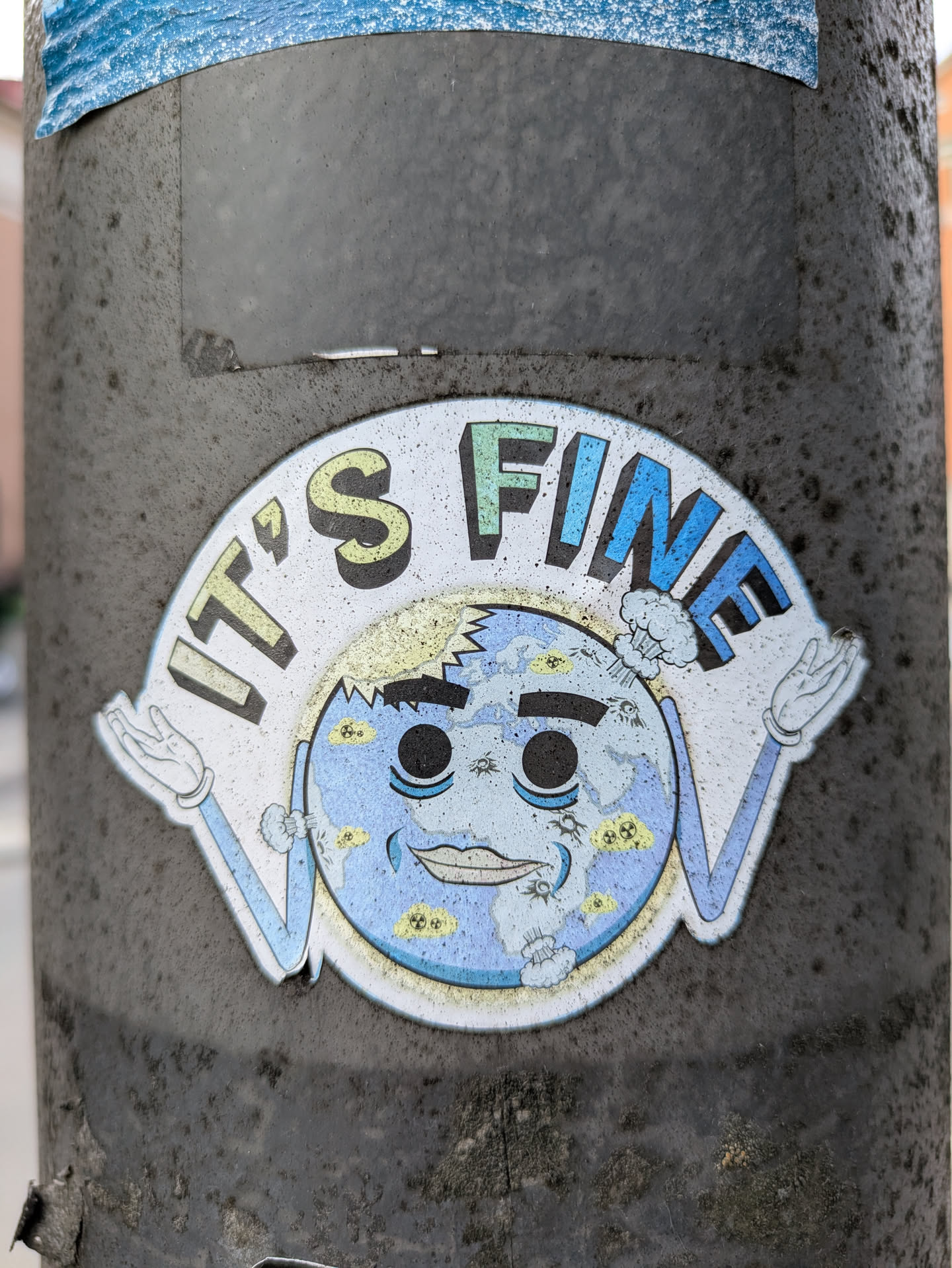 A round sticker on a grey post features a cartoon Earth with arms and a face. The Earth has a crack on its head, but it's shrugging and smiling. Text above reads, "It's fine."