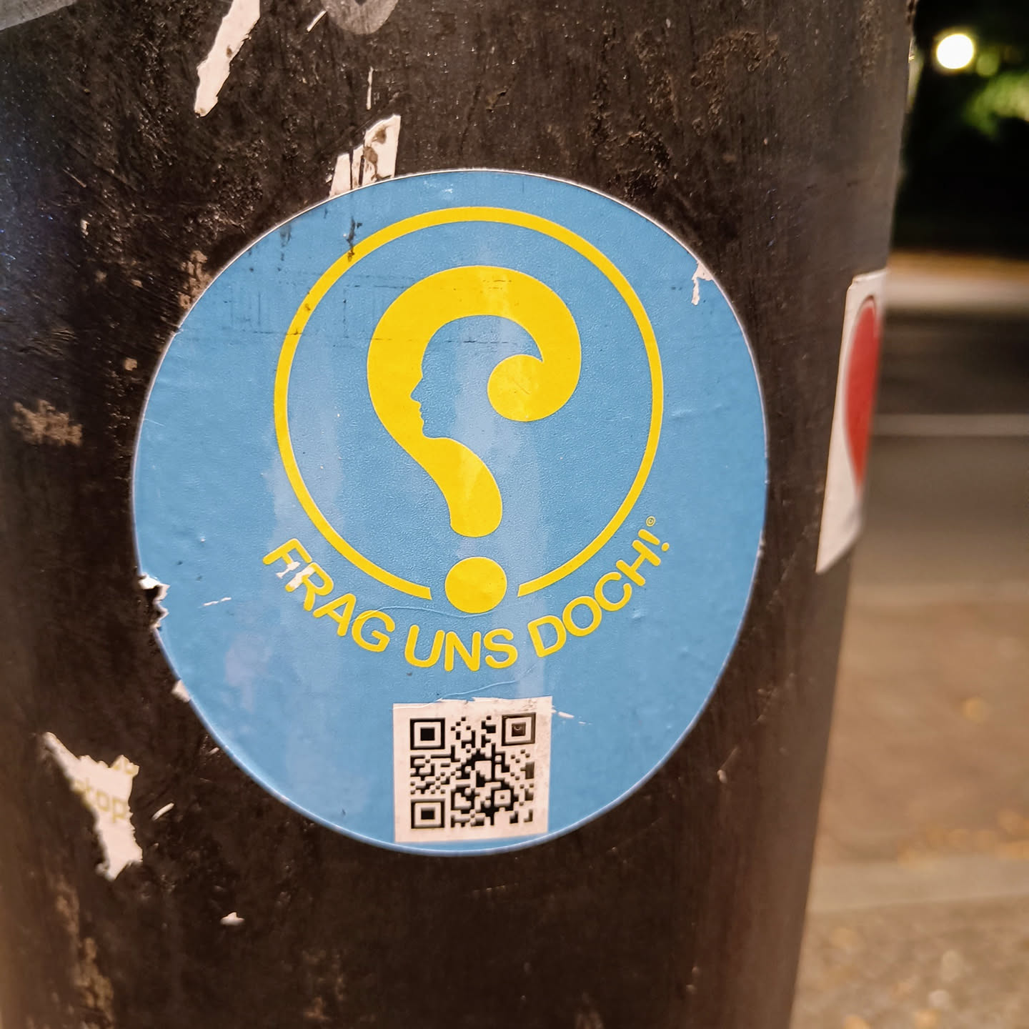 A light blue round sticker with a yellow question mark that includes a profile.  The text "FRAGE UNS DOCH!" is visible below the question mark. A QR code is located at the bottom of the sticker. The sticker is stuck on a dark-colored pole.