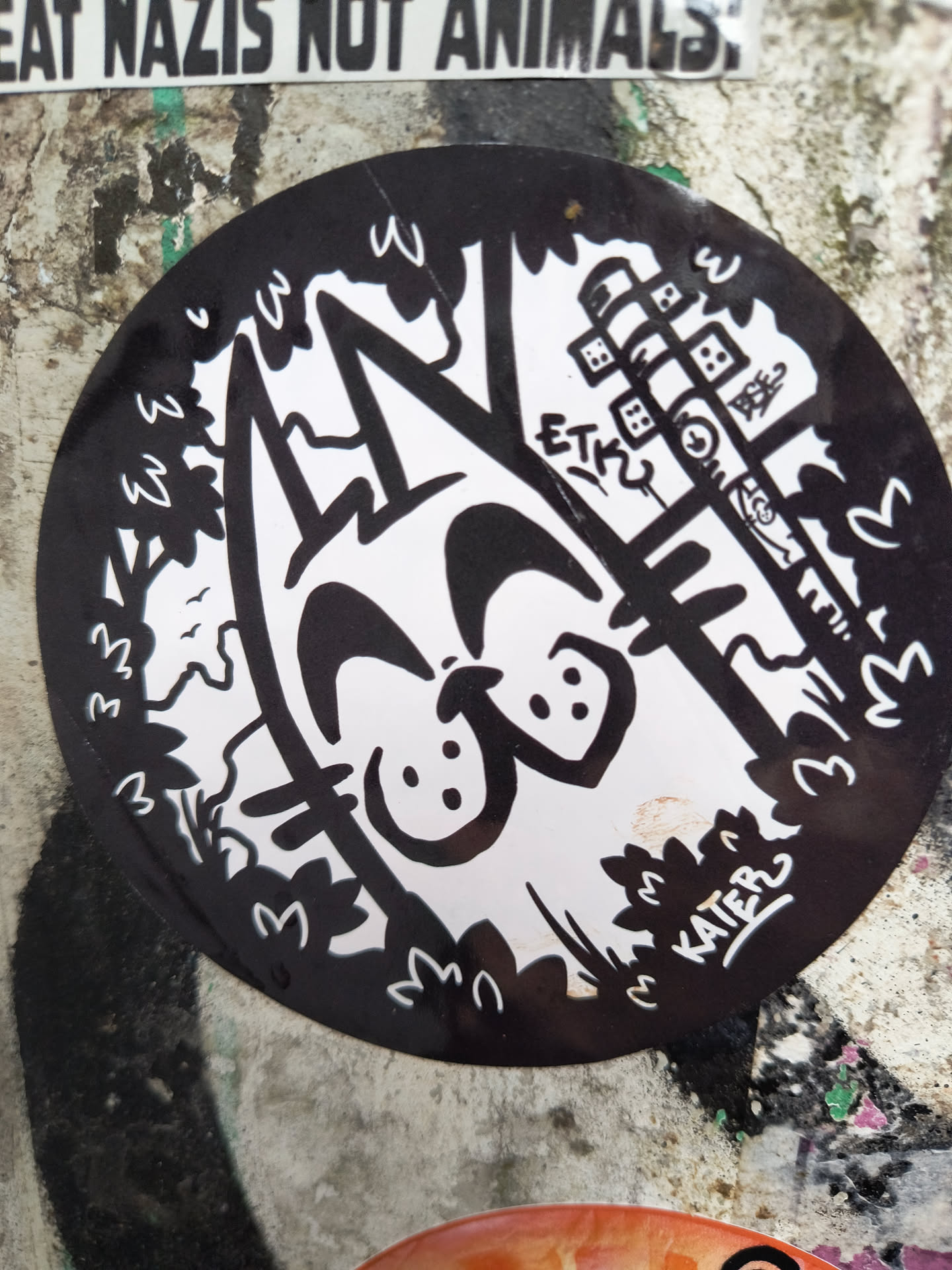 The sticker is circular and features a high-contrast black and white illustration. At its center is a stylized, smiling cat's face with large eyes and a wide, upturned mouth, reminiscent of a cartoon character. The cat's body is partially visible, blending into the surrounding elements. The background is a mix of organic, wavy shapes representing foliage and trees on the left and bottom, and more geometric, tower-like structures with small windows or blocks on the right side. Small 'W' shapes are integrated into the tree lines. The overall style is playful and somewhat abstract, with clean lines forming the white elements against a solid black background.
