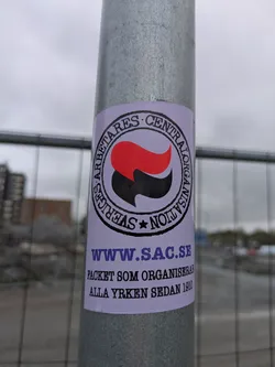 A sticker on a gray pole features the logo of Sveriges Arbetares Centralorganisation (SAC), a Swedish anarcho-syndicalist union. The logo contains a red and black flag inside a circle, with the union's name written around it. Below the logo, the text "www.sac.se" is displayed along with the text "PACKET SOM ORGANISERAR ALLA YRKEN SEDAN 1910".