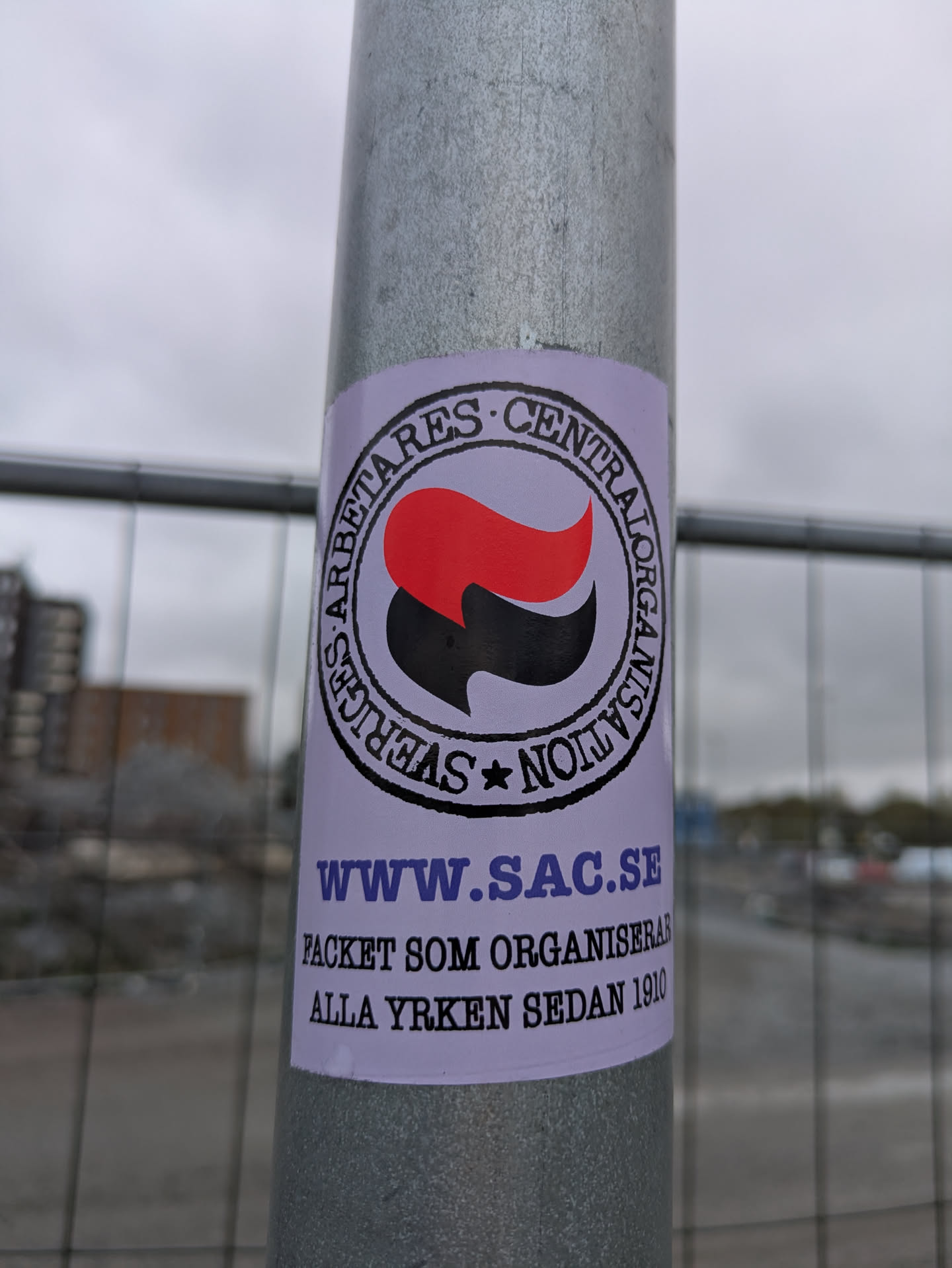A sticker on a gray pole features the logo of Sveriges Arbetares Centralorganisation (SAC), a Swedish anarcho-syndicalist union. The logo contains a red and black flag inside a circle, with the union's name written around it. Below the logo, the text "www.sac.se" is displayed along with the text "PACKET SOM ORGANISERAR ALLA YRKEN SEDAN 1910".