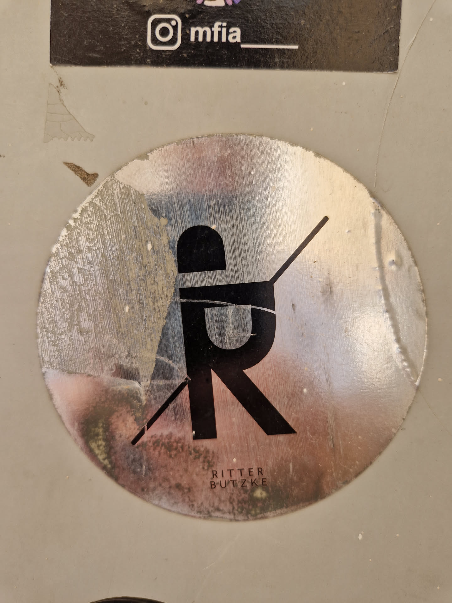 A round, silver sticker with a minimalist black logo featuring the letters 'R' and 'B' intersecting.  The text 'Ritter Butzke' is visible below the logo. The sticker shows some signs of wear and tear, adding to its unique character. An Instagram handle (@mfia) is visible on a sticker above the main sticker.