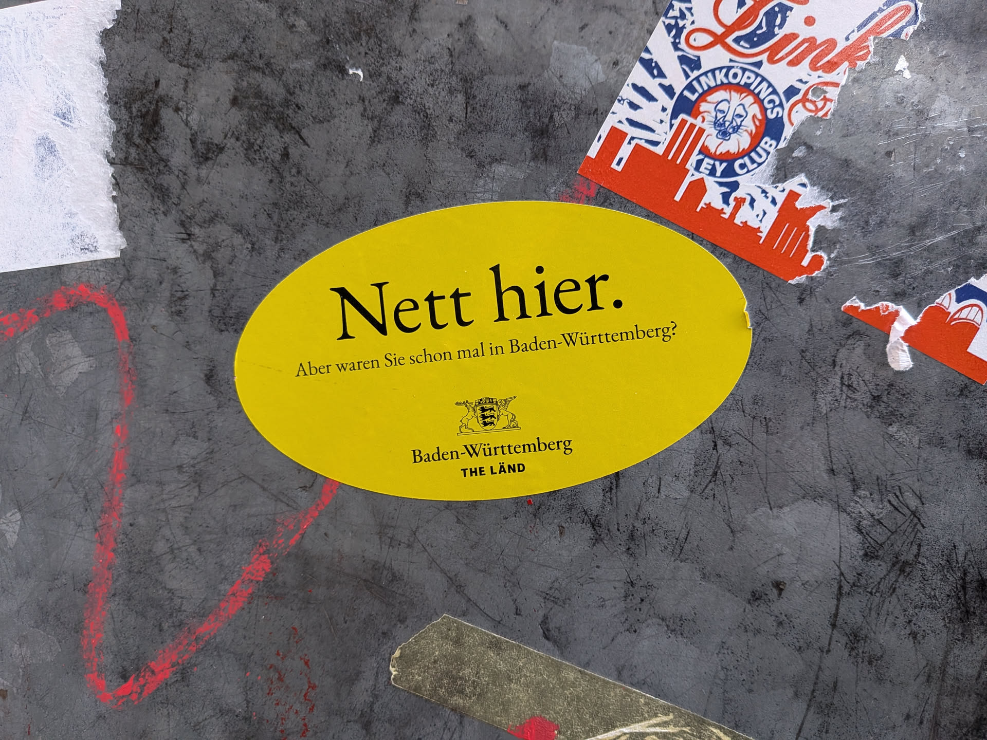 An oval-shaped, bright yellow sticker adhered to a scratched metallic surface. It displays black text reading 'Nett hier.' in a large serif typeface, followed underneath by the question 'Aber waren Sie schon mal in Baden-Württemberg?'. Near the bottom center is the coat of arms of the German state of Baden-Württemberg, above the state name and the official promotional campaign slogan 'THE LÄND'.