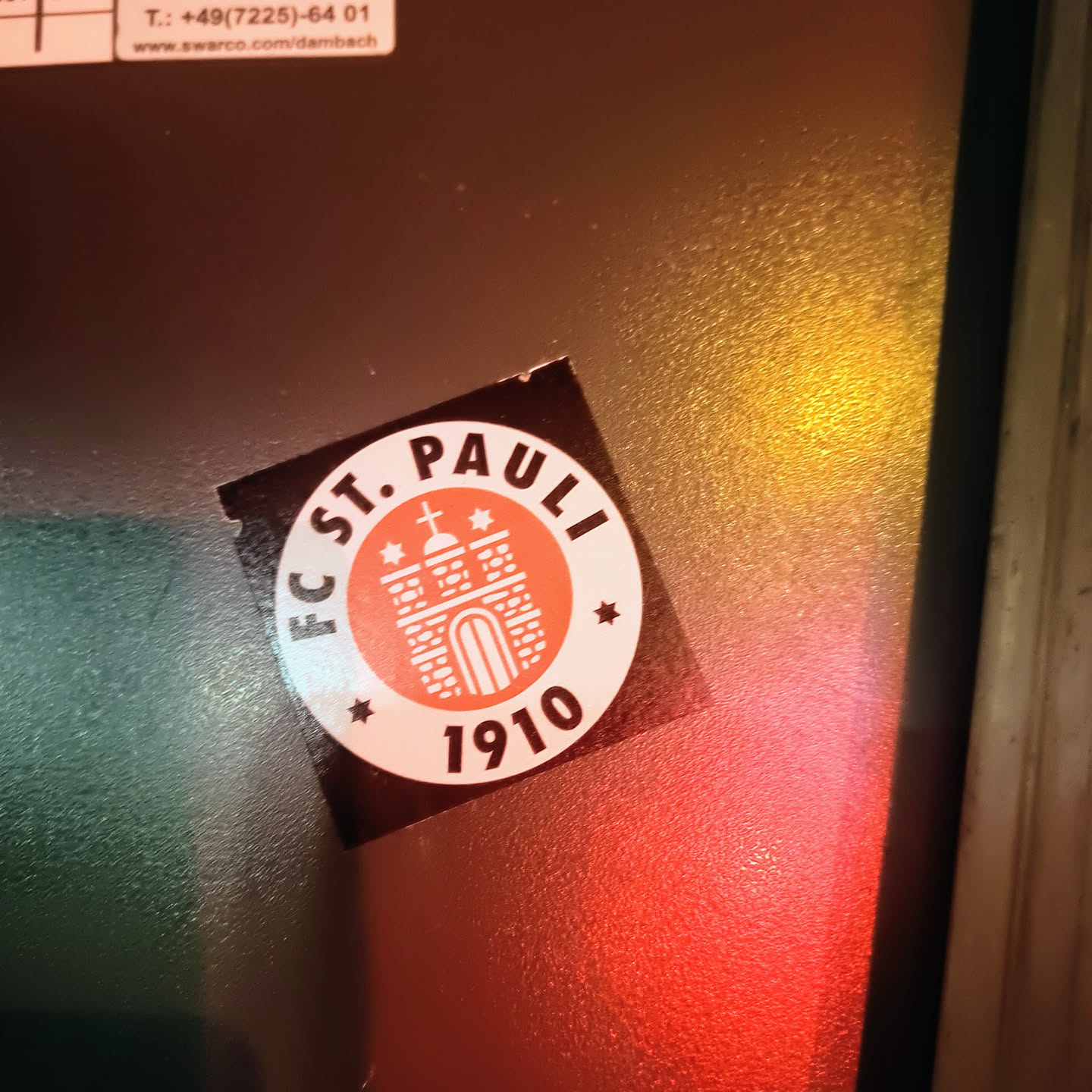 A round sticker featuring the logo of FC St. Pauli, a German football club. The logo depicts a stylized gate with three towers and a cross on top, set within a circular border with the club's name and founding year (1910). The sticker is dark-colored with the logo in shades of orange and white.