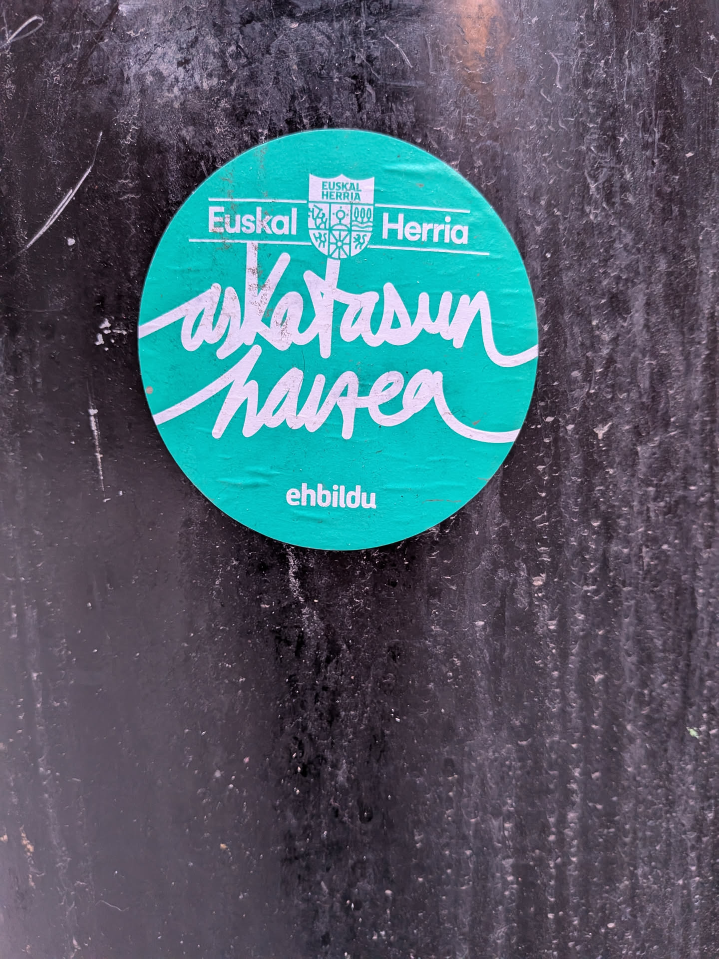 A circular sticker with a teal background and white text. At the top, there's a small crest-like emblem with 'EUSKAL HERRIA' written above it. Below the crest, 'Euskal Herria' is written in a sans-serif font. The dominant feature is the large, flowing cursive text in the center that reads 'askatasun haizea'. At the bottom, in a smaller sans-serif font, is the word 'ehbildu'. The sticker shows signs of wear and slight creasing, indicating it's adhered to a textured, dark surface.
