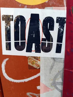 A minimalist sticker with the word TOAST in bold, uppercase, black letters on a white background. The sticker shows some wear and tear, suggesting it's been exposed to the elements. The word 'TOAST' is simple and impactful, and the design is very clean and modern. The sticker is affixed to an orange surface, which is itself covered in other layers of stickers, paint, and markings.