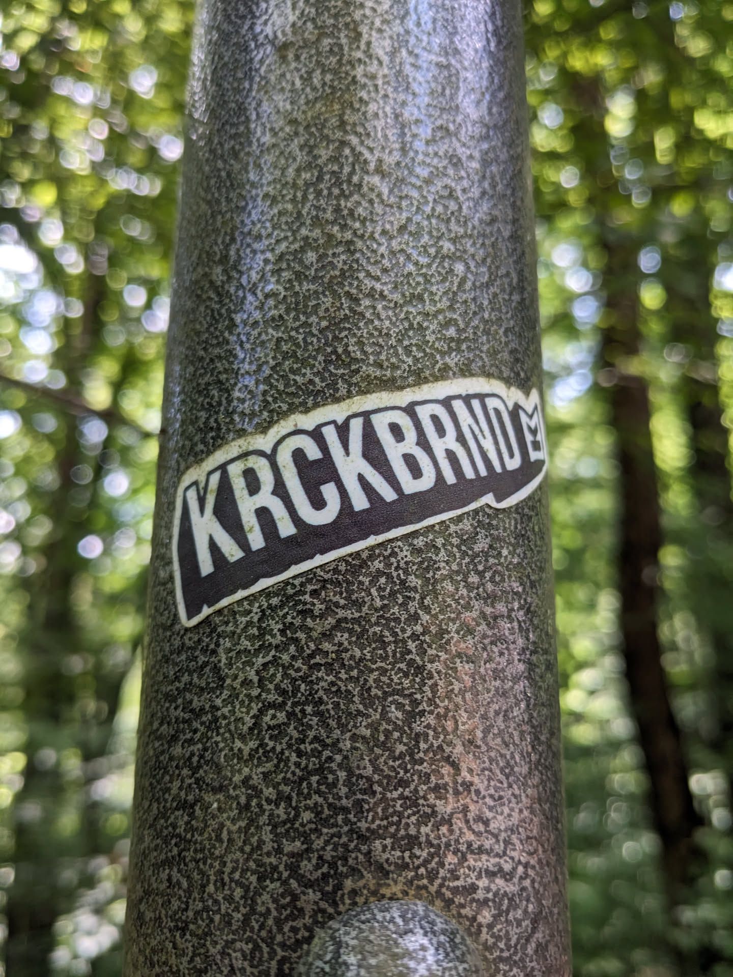 A dark gray and white sticker with the text 'KRCKBRND' is affixed to a textured gray pole in a forest setting. The sticker has a slightly curved rectangular shape with a small symbol near the end of the text. The background is blurred and shows the green foliage of the trees.
