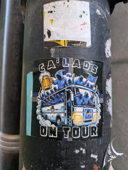 A square-format sticker featuring an illustration of a bus filled and surrounded by anonymous figures clad in blue hooded jackets. One hooded figure standing on top holds a large frothy mug of beer, while others wave blue-and-white flags and pump their fists. The bus is rendered in light blue and white with illuminated headlights. Above the illustration, stylized distressed lettering spells 'CA LADS', and below it, 'ON TOUR'. The background is primarily dark with a horizontal blue and grey accent stripe.