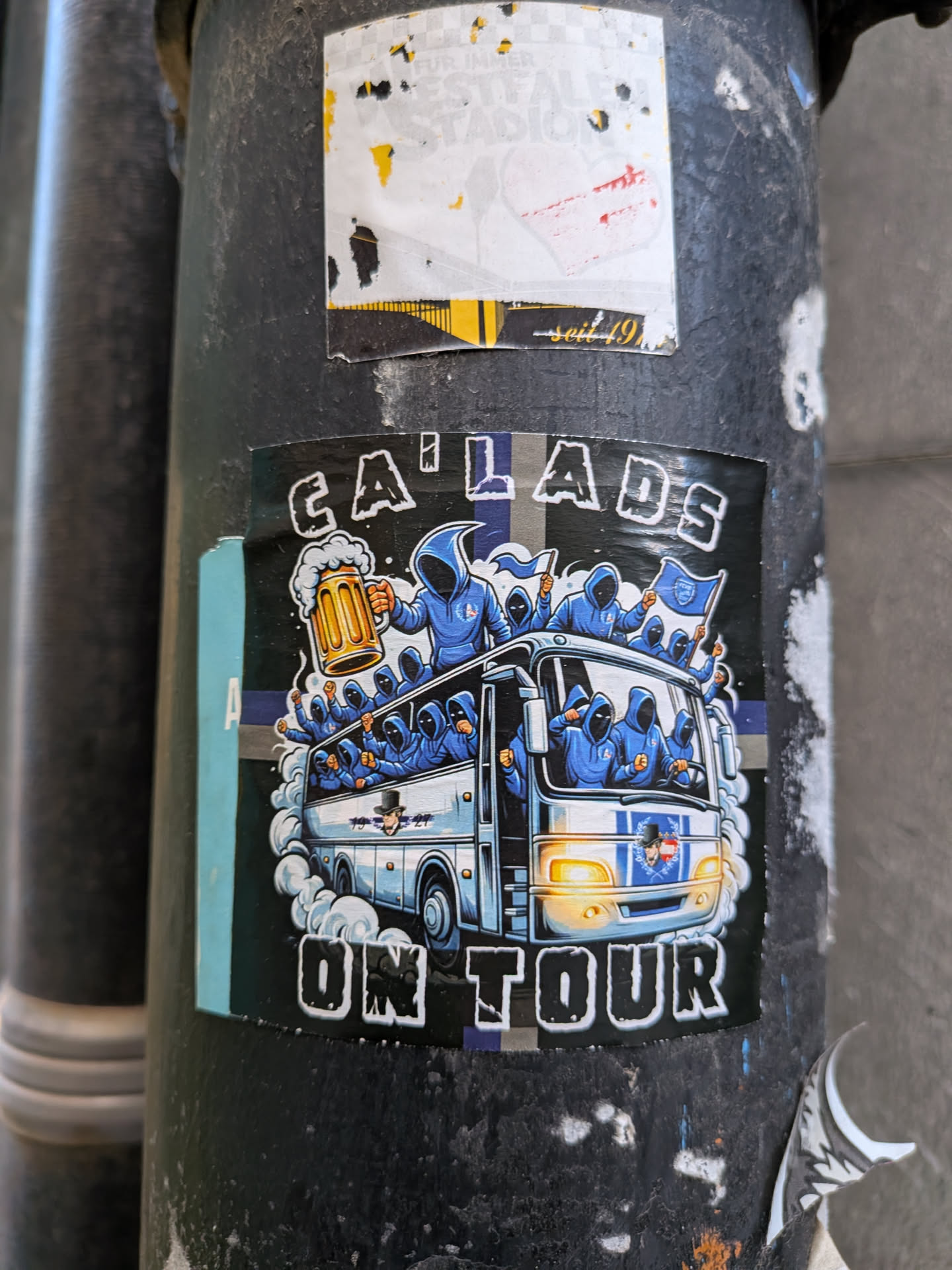 A square-format sticker featuring an illustration of a bus filled and surrounded by anonymous figures clad in blue hooded jackets. One hooded figure standing on top holds a large frothy mug of beer, while others wave blue-and-white flags and pump their fists. The bus is rendered in light blue and white with illuminated headlights. Above the illustration, stylized distressed lettering spells 'CA LADS', and below it, 'ON TOUR'. The background is primarily dark with a horizontal blue and grey accent stripe.