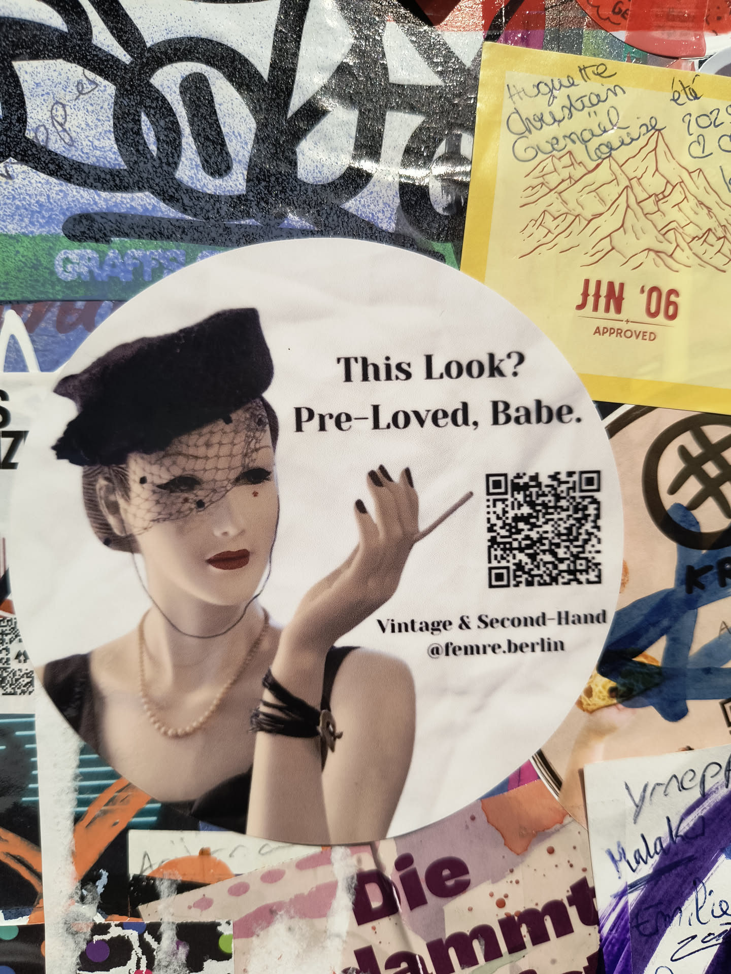 A circular sticker featuring a stylized image of a mannequin or model wearing a black cloche hat with a veil, a pearl necklace, and a black dress. The model has red lipstick and is holding a cigarette. To the right of the model is a QR code. Below the QR code, the text 'Vintage & Second-Hand @femre.berlin' is visible. The sticker has a slightly aged appearance.