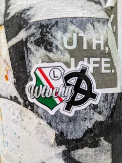 A sticker is affixed to a pole. It has a shield design with the colors of the Italian flag, the letter "L", and the word "Wlachy" written in a stylized script. A black symbol is also present on the sticker. The background shows other torn posters and stickers, indicating an urban setting.