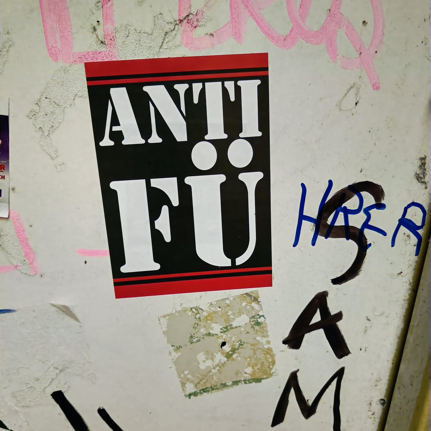 A black and red sticker with the text "ANTI FU" in white stencil lettering. The sticker is affixed to a wall with other graffiti and stickers.