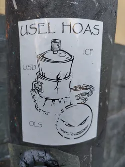 Black and white sticker with an illustration of a chained spray can and a grinning face. The text 'USEL HOAS' is at the top, with additional markings 'USD', 'ICF', and 'OLS' on the sticker. The style is reminiscent of graffiti art.